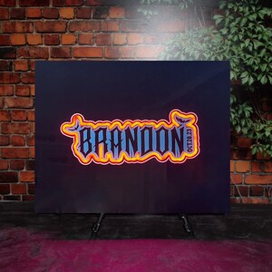 Op de afbeelding: Een zwart bord met een neonoranje en gele omtrek van het woord "BRANDON" met een vleermuisafbeelding en de datum "OCT 28 23".