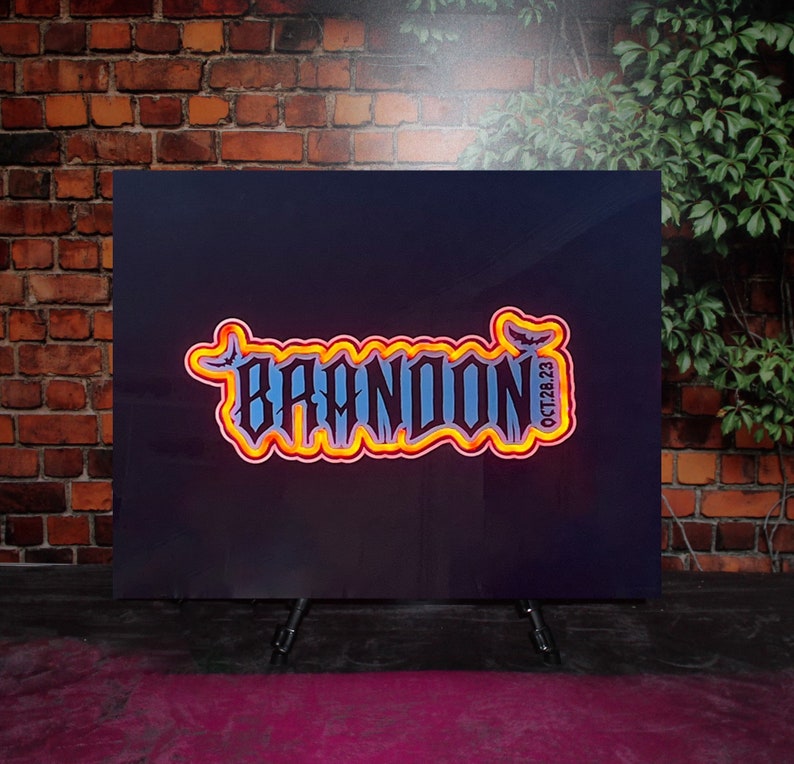 Op de afbeelding: Een zwart bord met een neonoranje en gele omtrek van het woord "BRANDON" met een vleermuisafbeelding en de datum "OCT 28 23".