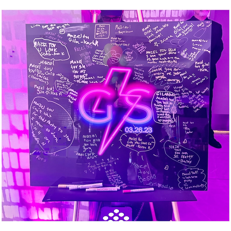 Op de afbeelding: Een glazen bord met een neonroze en blauwe bliksemgrafiek met de letters "GS" en de datum "03.26.23". Het bord is bedekt met handgeschreven berichten in zwarte stift, die iemand geluk wensen en feliciteren. De berichten omvatten "Mazel Tov", "Je ziet er geweldig uit" en "Veel geluk met de test".