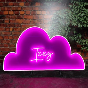 Op de afbeelding: Een roze neon-bord in de vorm van een wolk met de naam "Izzy" in wit neon erin. Het bord staat op een roze oppervlak voor een bakstenen muur.