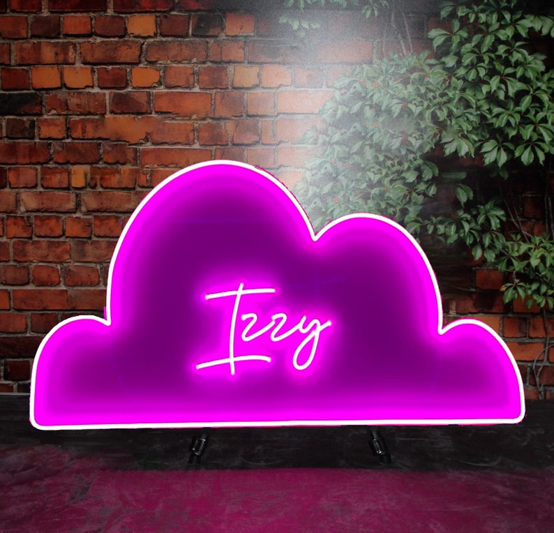 Op de afbeelding: Een roze neon-bord in de vorm van een wolk met de naam "Izzy" in wit neon erin. Het bord staat op een roze oppervlak voor een bakstenen muur.