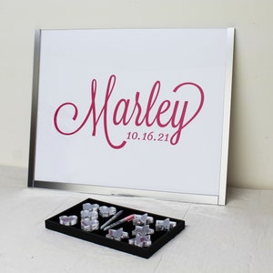 May include: A white framed sign with a pink inscription that reads "Marley 10.16.21".  A black tray with clear plastic heart-shaped containers and silver pens.