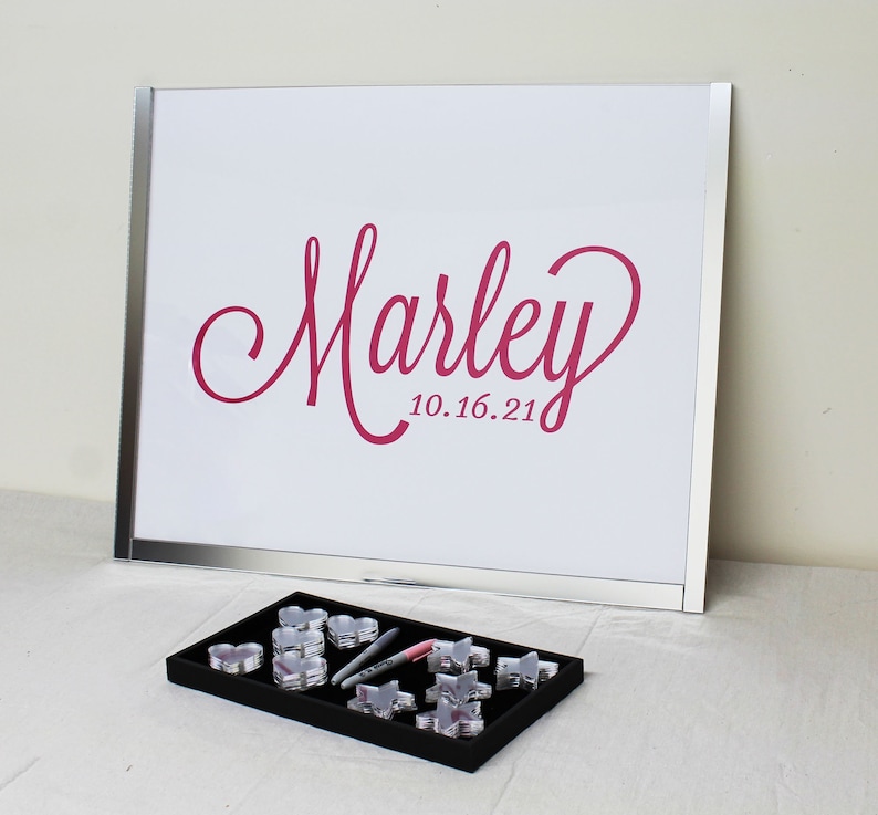 May include: A white framed sign with a pink inscription that reads "Marley 10.16.21".  A black tray with clear plastic heart-shaped containers and silver pens.