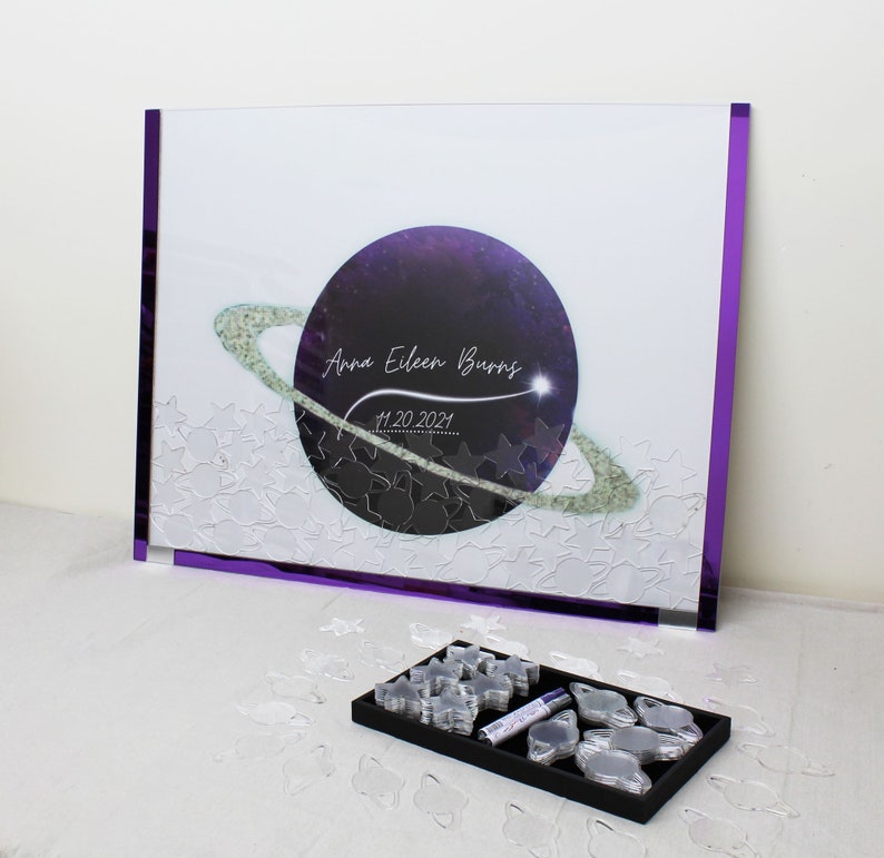 May include: A purple and white galaxy themed guest book with a clear plastic frame. The book has a purple planet with a ring around it and the text "Anna Eileen Burns 11.20.2021". The book comes with a set of clear plastic stars and planets for guests to sign.