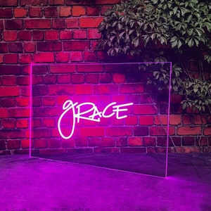 Op de afbeelding: Een neonbord met het woord "GRACE" in roze cursief schrift. Het bord is gemonteerd op een bakstenen muur en straalt een roze gloed uit.