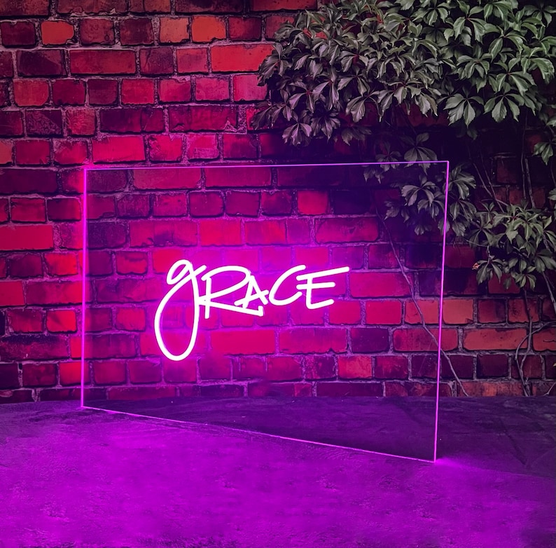 Op de afbeelding: Een neonbord met het woord "GRACE" in roze cursief schrift. Het bord is gemonteerd op een bakstenen muur en straalt een roze gloed uit.