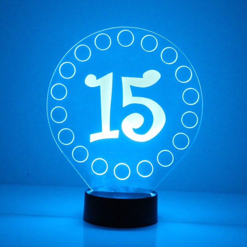 May include: A white acrylic night light shaped like a circle with a white number "15" in the center. The circle is surrounded by a ring of small circles.