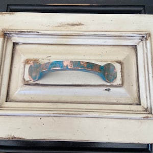 May include: A white drawer with a distressed finish and a copper-colored drawer pull with a teal blue patina.