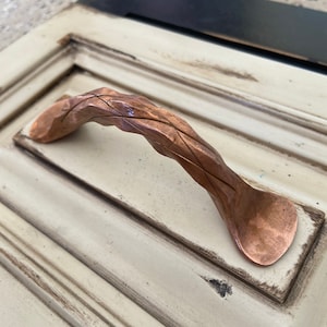 May include: A copper cabinet door handle with a woven design. The handle is curved and has a rustic finish.