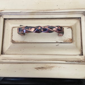 Op de afbeelding: Een gedraaide, koperkleurige ladegreep met een zwarte afwerking. De greep is gemonteerd op een witte kastdeur.