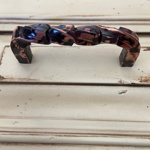 May include: A twisted, copper-colored cabinet pull with a black finish. The pull is designed to be used on a cabinet door or drawer.