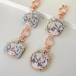 May include: Four keychains featuring anime-style character heads with light grey hair and blue eyes. Two are clear acrylic charms showing a character sleeping on a pillow, and two are wooden charms with rose gold-coloured bunny-shaped clasps.