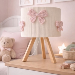 Könnte beinhalten: Eine cremefarbene Lampe mit zylindrischem Schirm und Holz-Dreibein-Sockel. Der Schirm ist mit drei gehäkelten rosa Schleifen verziert. Die Lampe steht auf einem Nachttisch aus Holz in einem Kinderzimmer, mit gerahmten Tierbildern an der Wand.