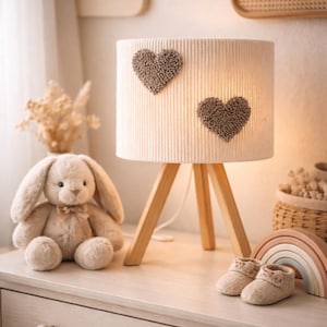 Könnte beinhalten: Eine neutral gehaltene Szene im Kinderzimmer mit einer cremefarbenen Lampe mit zwei Herzapplikationen, einem Plüschhasen, Babyschuhen und einem Holzregenbogen. Die Lampe hat ein Holzstativ. Dekorative Holzwolken an der Wand.
