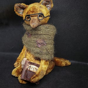 May include: A brown teddy bear wearing round glasses and a green knitted scarf, holding a brown book with the word "BOOK" written on it.