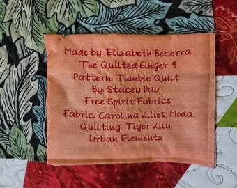 May include: A close-up of a peach-colored fabric label with red embroidered text. The text reads: "Made by Elizabeth Becerra The Quilted Ginger 9 Pattern: Tumble Quilt By Stacey Day, Free Spirit Fabrics Fabric: Carolina Lillies, Moda Quilting: Tiger Lilly, Urban Elements"