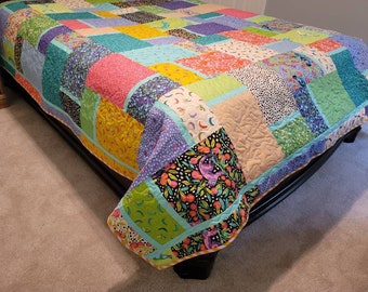 May include: A colourful patchwork quilt with a variety of floral and geometric patterns in shades of pink, green, yellow, blue, and purple. The quilt is made of different fabrics and is laid out on a bed.