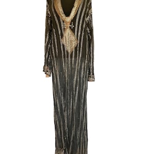 Puede incluir: Un vestido largo negro con rayas verticales doradas punteadas y adornos dorados. El vestido presenta un escote en V y un diseño en forma de diamante en el pecho. Las mangas son largas y el dobladillo tiene un borde con volantes. El vestido se exhibe en un maniquí negro.