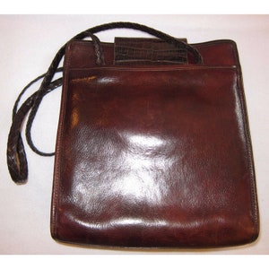 Pu&ograve; includere: Una borsa a mano in pelle marrone con una tracolla intrecciata e una patta con effetto coccodrillo.