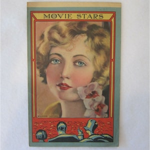 May include: A vintage movie star card featuring a portrait of Marion Davies, a blonde woman with blue eyes, wearing a white dress and holding a white orchid. The card has a red and blue border and the text "MOVIE STARS" at the top.