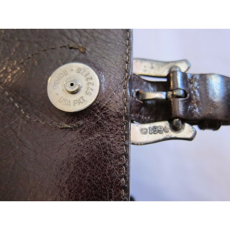 Pu&ograve; includere: Primo piano di una borsa di pelle marrone con una fibbia argentata e un'etichetta argentata che recita "5722125 ROMAG USA PAT". La fibbia ha l'anno "1996" inciso.