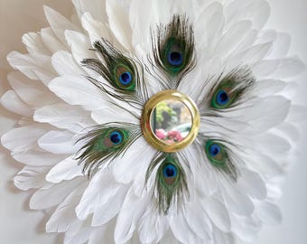May include: A decorative wall mirror featuring a circular design of white feathers. The center has a small round mirror framed in gold, surrounded by peacock feathers with blue and green accents. The overall shape resembles a flower.