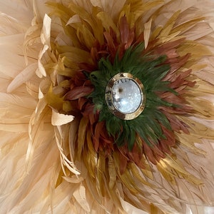 Peut inclure: Une décoration murale ronde en plumes de couleur beige, marron et verte. Au centre de la décoration se trouve un petit miroir rond avec un cadre doré.