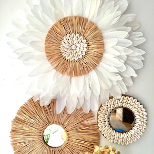 Peut inclure: Décor mural composé d'une grande fleur en plumes blanches, de deux miroirs ronds et d'un petit miroir. Les miroirs sont encadrés de matières naturelles et de coquillages. L'ensemble est disposé sur un mur blanc.