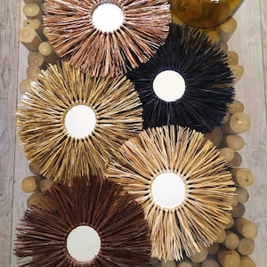 Peut inclure: Cinq miroirs muraux ronds avec un cadre en paille tressée. Les miroirs sont dans différentes nuances de brun, noir et paille naturelle. Les miroirs sont disposés en cercle sur une surface en bois.