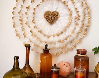 May include: A circular wall hanging composed of layered white feathers with gold-tipped edges, featuring a central heart-shaped cluster of small gold beads. The piece is displayed above an assortment of amber glass bottles and a pink salt lamp.