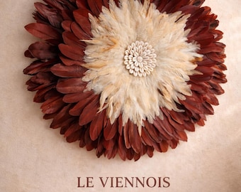 May include: Circular wall decor made of layered feathers in shades of burgundy and cream, with a central design of small white shells. The text "LE VIENNOIS" is displayed below the decor. The piece is mounted on a beige wall.