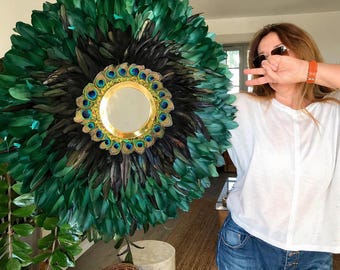 May include: A round wall mirror with a gold frame and a peacock feather design. The mirror is surrounded by green and black feathers, with a gold and blue peacock feather border around the mirror's edge. The mirror is a decorative wall hanging.