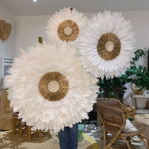 Puede incluir: Tres grandes piezas de decoración de pared de plumas blancas con detalles dorados. Las piezas están dispuestas en forma escalonada en una pared blanca.
