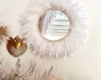 May include: A round mirror with a white feather frame and a gold beaded border. The mirror is reflecting a white wall with a window.