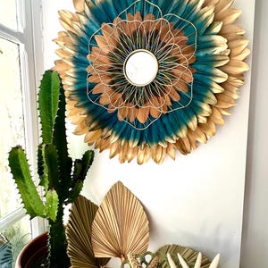 Puede incluir: Un espejo de pared decorativo con un diseño circular hecho de plumas en tonos turquesa, dorado y marrón. El espejo tiene un marco de alambre dorado y está montado en una pared blanca. Un cactus en una maceta estampada y objetos decorativos están en primer plano.