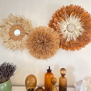 Peut inclure: Trois décorations murales circulaires aux textures et couleurs naturelles. Les décorations ressemblent à des fleurs, faites de plumes et de matériaux tissés. En dessous, diverses bouteilles et vases en verre ambré sont disposés sur une surface blanche, créant une esthétique bohème.