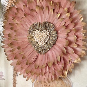 Peut inclure: Décoration murale circulaire composée de plumes roses avec des pointes dorées. Le centre présente un cœur fait de petits coquillages blancs. L'ensemble est monté sur un fond neutre, créant une esthétique douce et élégante.