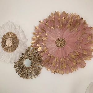 May include: Three wall decor pieces in a bohemian style. One is a large pink and gold feather flower, one is a smaller white feather flower, and the third is a round, gold, woven piece with a mirror in the center.