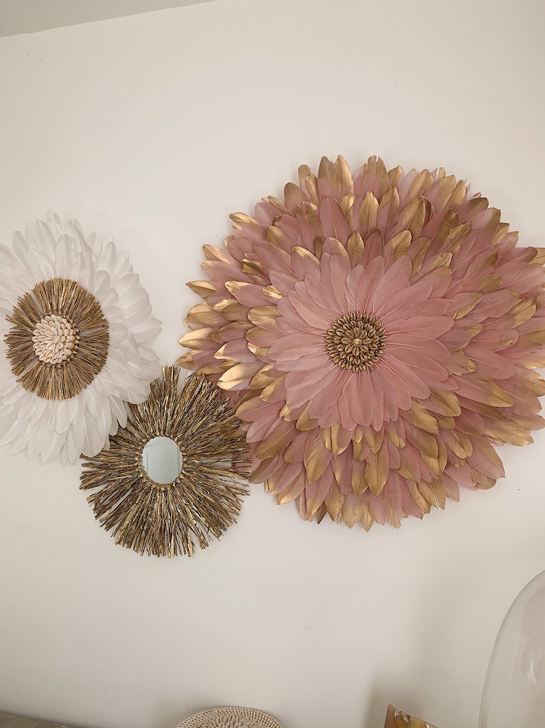 May include: Three wall decor pieces in a bohemian style. One is a large pink and gold feather flower, one is a smaller white feather flower, and the third is a round, gold, woven piece with a mirror in the center.