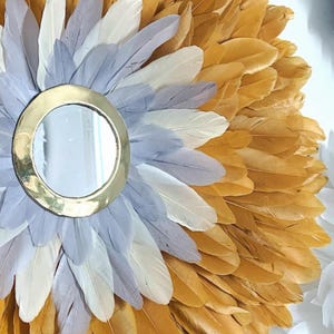 May include: A round mirror with a gold frame surrounded by a wreath of feathers. The feathers are in shades of gold, white, and gray.