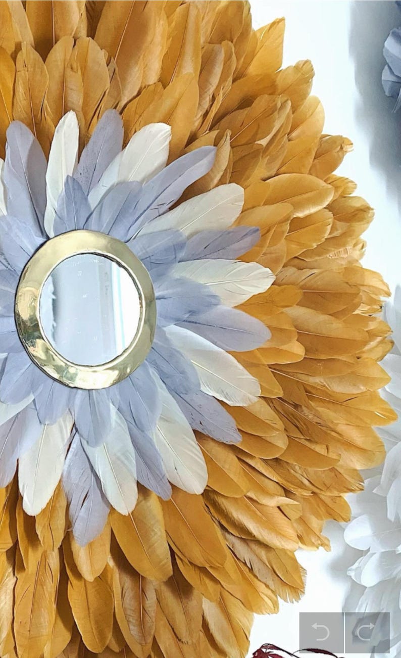 May include: A round mirror with a gold frame surrounded by a wreath of feathers. The feathers are in shades of gold, white, and gray.