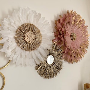 May include: Three wall decor pieces featuring a large white feather flower, a smaller pink feather flower, and a round gold mirror with a woven frame.