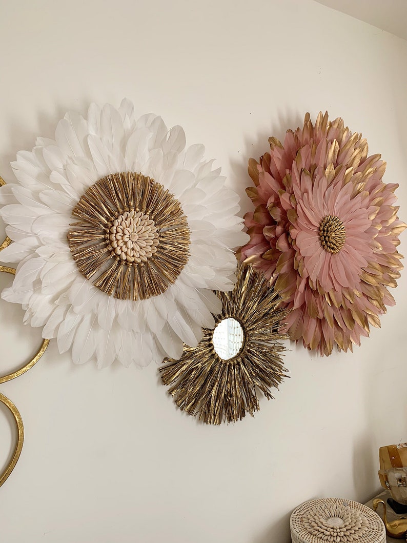 May include: Three wall decor pieces featuring a large white feather flower, a smaller pink feather flower, and a round gold mirror with a woven frame.