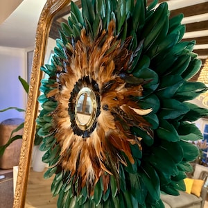 May include: A round mirror with a green and brown feather wreath. The mirror is surrounded by a gold frame.