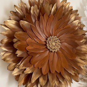 Peut inclure: Une grande décoration murale en forme de fleur, faite de plumes marron et dorées. Le centre présente un groupe de petits coquillages dorés. Les plumes sont disposées en couches, créant un effet de dimension.