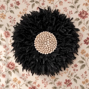 Peut inclure: Une décoration murale circulaire composée d'une couche dense de plumes noires rayonnant autour d'un centre formé de coquillages cauris blanc cassé disposés en motif concentrique, offrant un aspect texturé et superposé.