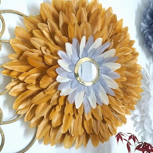 May include: A round wall mirror with a gold frame surrounded by a large circle of mustard yellow feathers. The center of the mirror is surrounded by a smaller circle of white and gray feathers.
