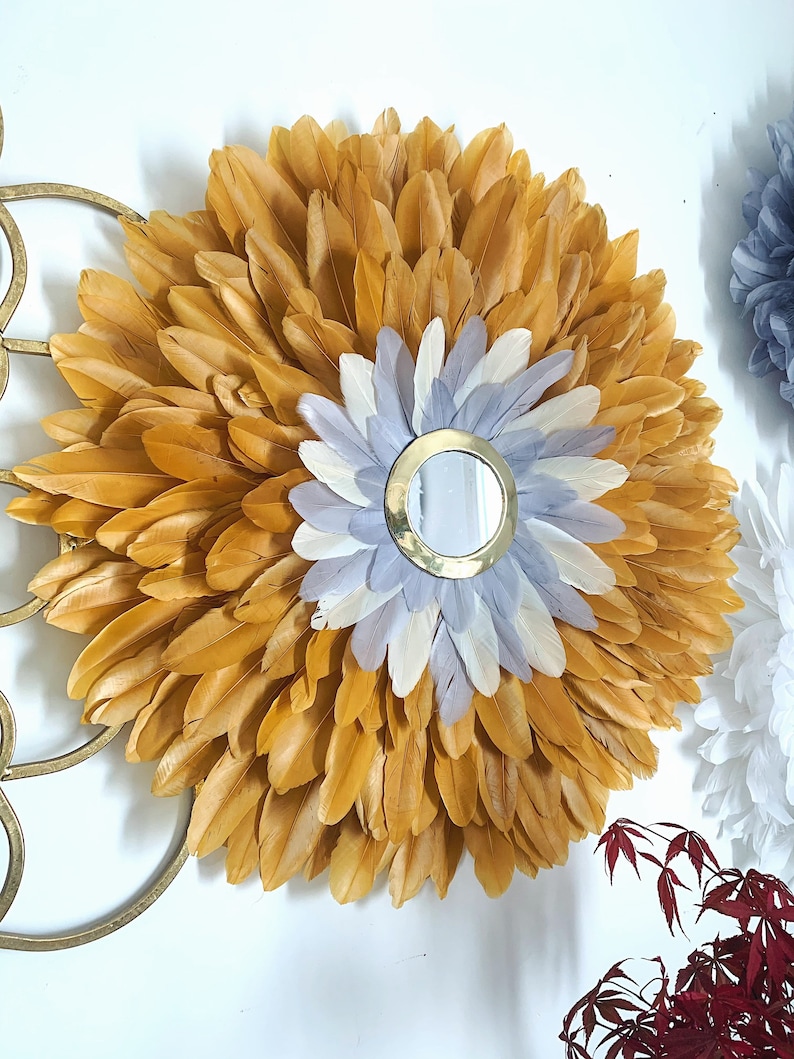 May include: A round wall mirror with a gold frame surrounded by a large circle of mustard yellow feathers. The center of the mirror is surrounded by a smaller circle of white and gray feathers.