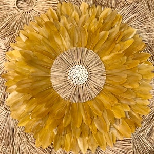 May include: A large, round wall hanging made of natural materials. The center of the hanging is a woven circle with a cluster of white seashells. The circle is surrounded by a ring of golden feathers.