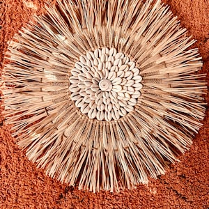 May include: A round wall decor made of natural materials. The center of the decor is a circle of white seashells. The outer edge of the decor is made of woven straw with long strands of straw extending outward.